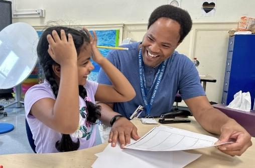  Christopher Lane smiles as he holds a piece of paper while sitting next to a student.